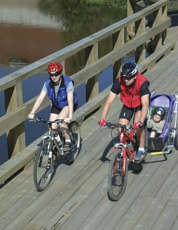 Deux adultes à vélo transportent un jeune enfant dans une remorque sur un pont du Petit Témis